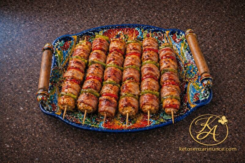 Spiedini di pollo avvolti nel bacon con verdure grigliate, serviti su un grande vassoio colorato con manici in legno, su sfondo marrone con logo ketosenzarinunce.com.