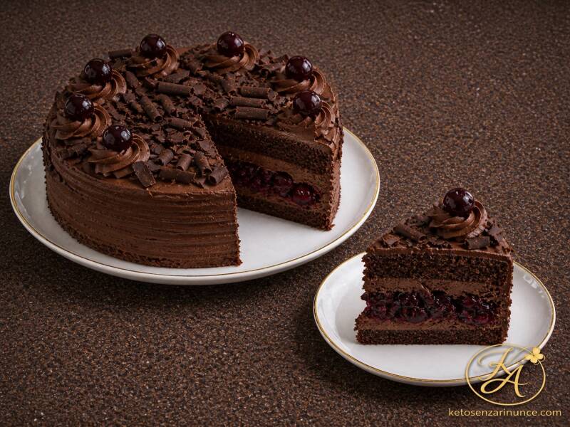 Torta chetogenica al cioccolato con rum e amarena, decorata con ganache al cioccolato, amarene e riccioli fondenti. Una fetta tagliata mostra il ripieno scuro e morbido di amarene.