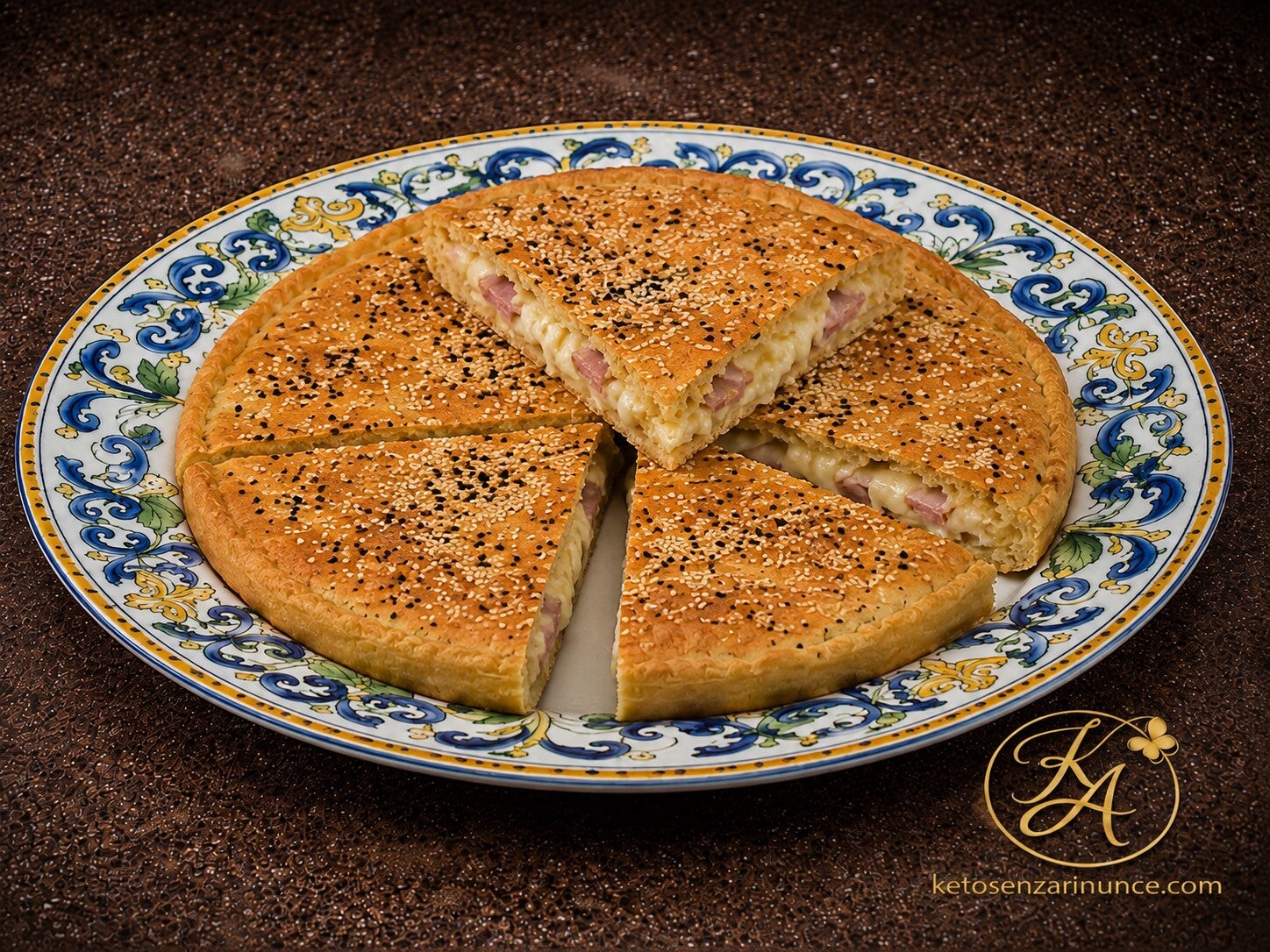Torta salata chetogenica ripiena con prosciutto cotto e mozzarella, dorata in superficie e decorata con semi di sesamo bianchi e neri, servita su un grande piatto con bordo mediterraneo colorato.