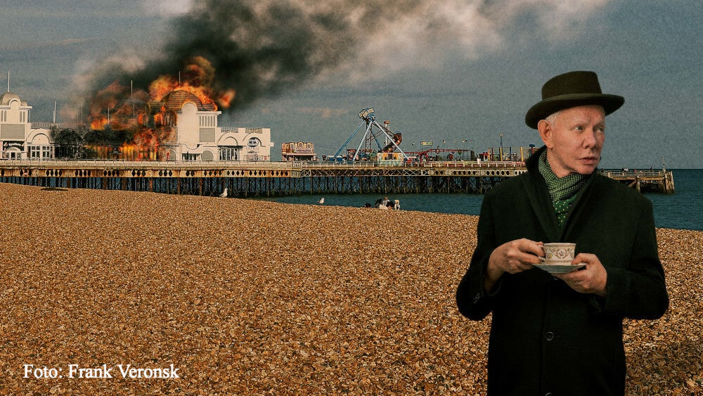 Joe Jackson mit Teetasse, Mantel und Hut am Strand, Hintergrund ein brennendes Kai-Gebäude