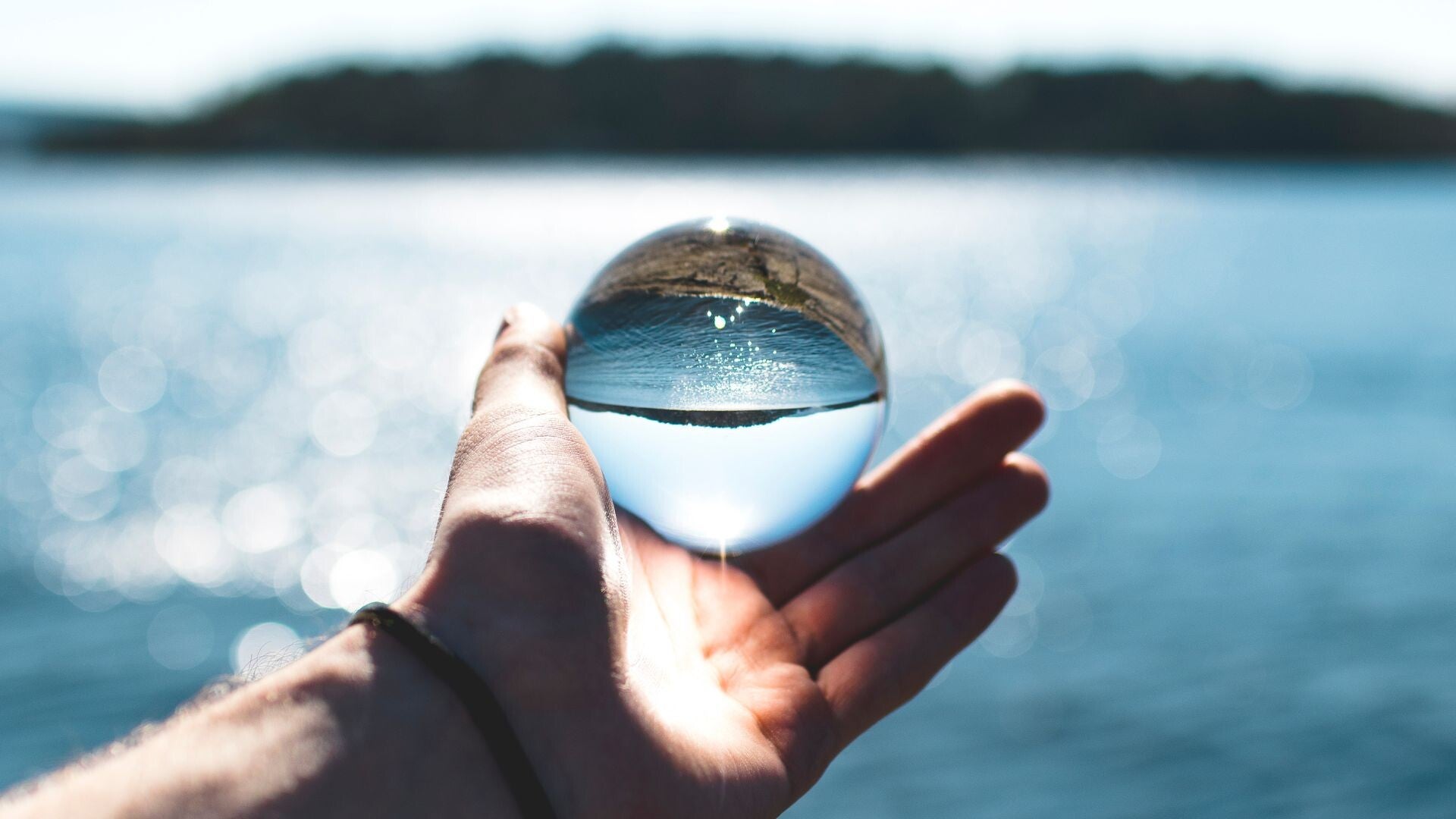 Hand hält eine Glaskugel, die spiegelt den klaren blauen See - Symbol für Reiniung, Klärung und Innere Transformation