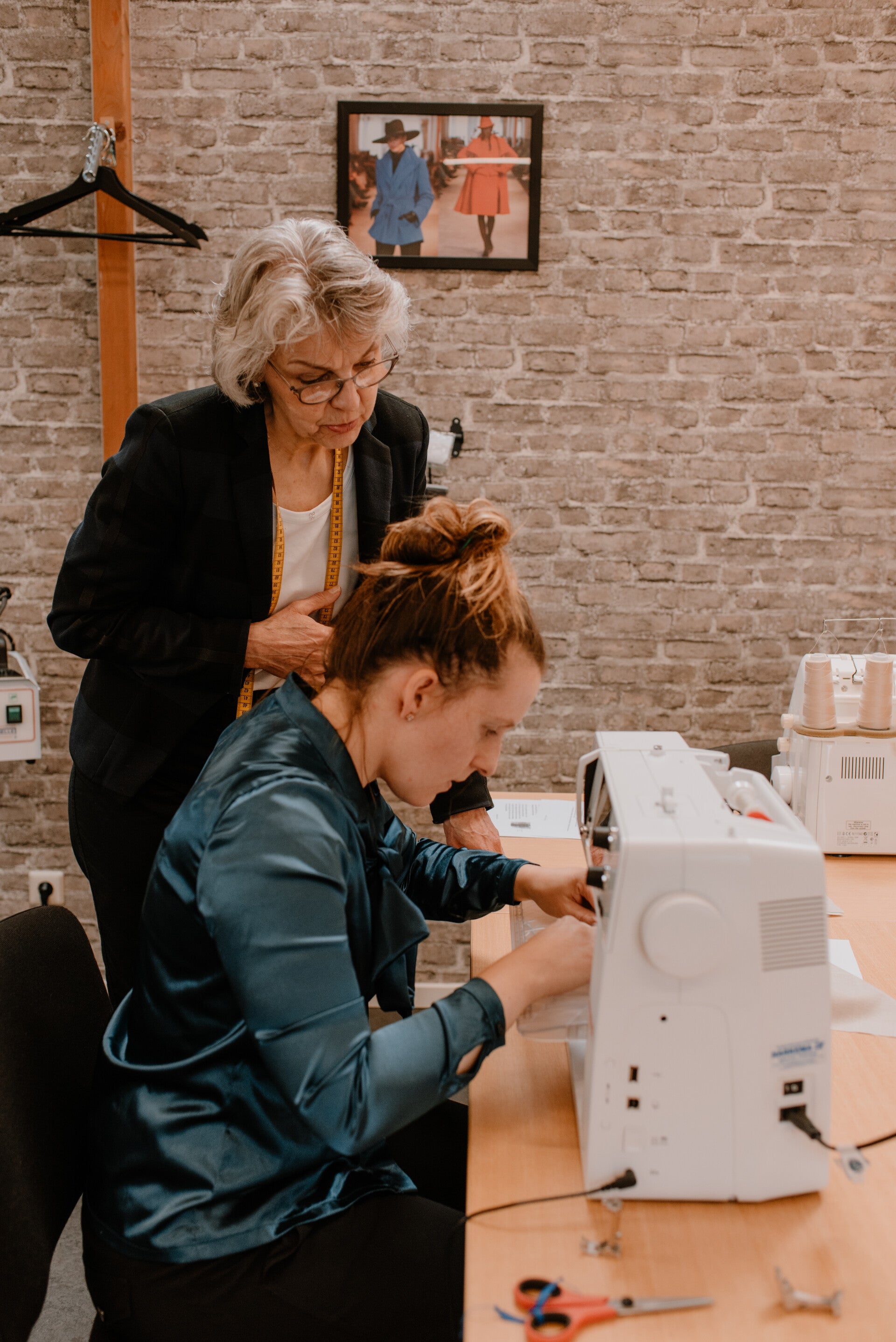 Sfeerimpressie naaicursus voor beginners bij Modestudio Jacoba in Groningen. Eigenaar en docent Coba Steenbergen kijkt mee met een cursist die zit te naaien. Coba draagt een zwart gestreepte blazer, de cursist een donkergroene blouse.