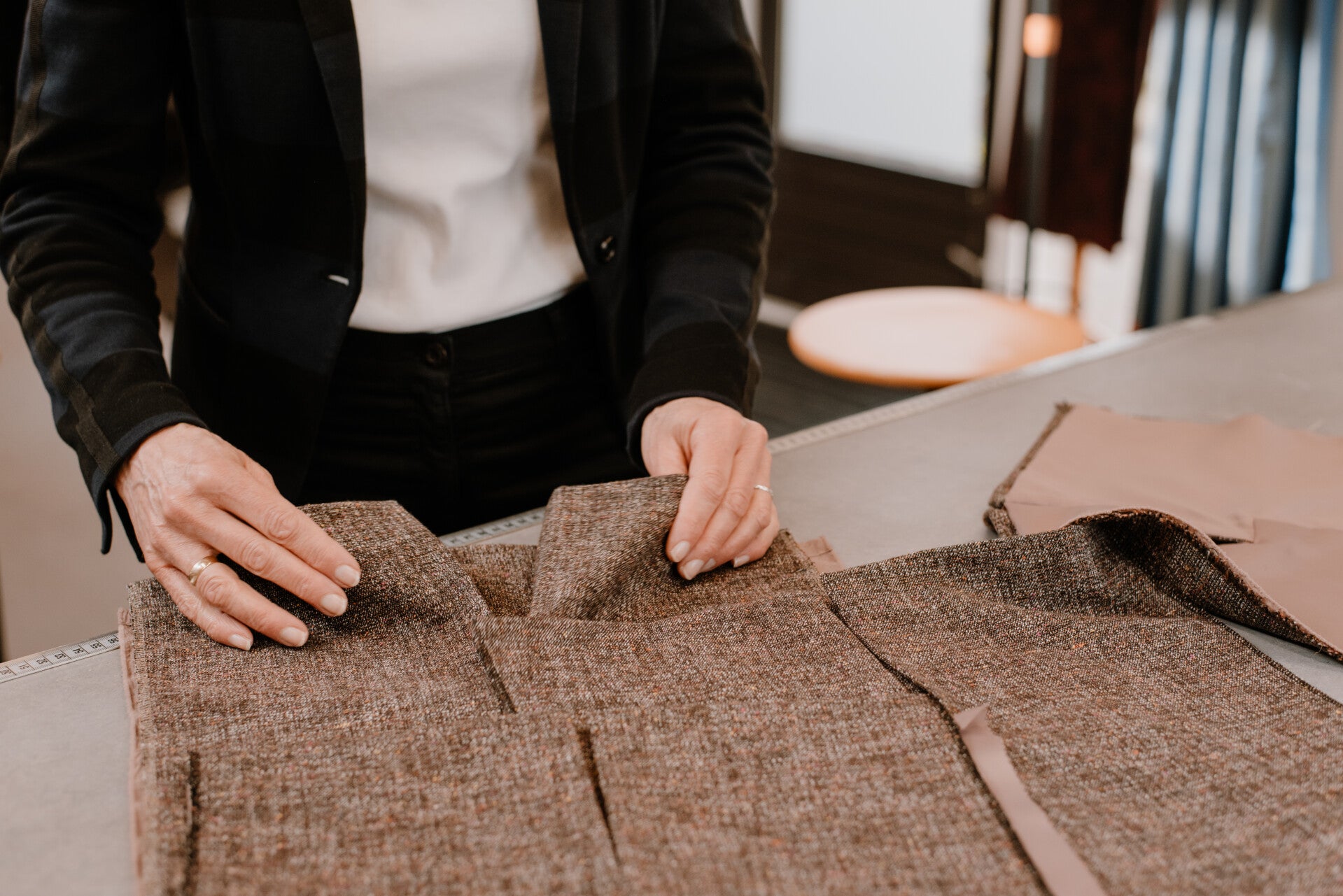 Sfeerimpressie van de opleiding tot coupeuse/kleermaker. Op tafel ligt bruine mantelstof, Coba Steenbergen heeft de stof in handen, ze draagt een zwarte gestreepte blazer.
