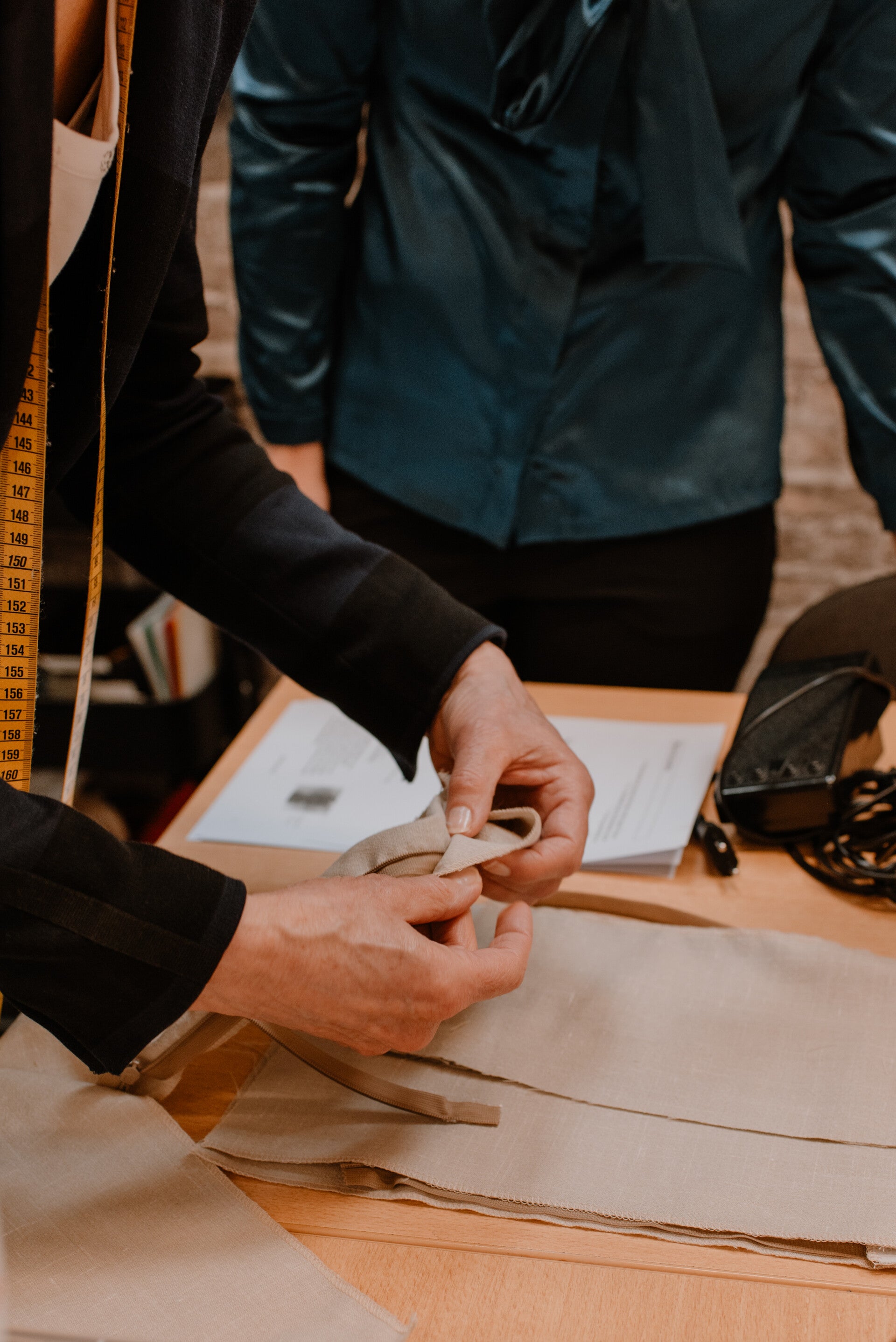 Sfeerimpressie van de modevakopleiding docent. Coba Steenbergen bekijkt het naaiwerk van een cursist. Ze draagt een zwart gestreepte blazer en heeft een geel meetlint om haar hals hangen. De cursist naast haar draagt een donkergroene blouse.