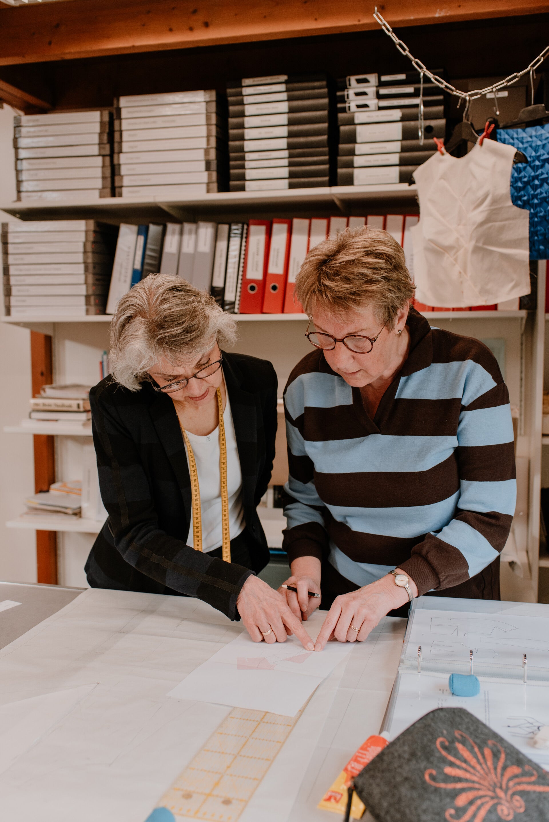 Sfeerimpressie van de naaicursus Basis Mode Maken. Coba Steenbergen draagt een zwart gestreepte blazer en heeft een geel kleermakerslint om haar hals hangen, ze helpt een cursist en wijst iets aan. De cursist draagt een blauw-zwart gestreepte trui.