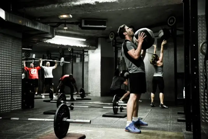 une salle de sport avec un homme en premier plan qui porte une wall ball et d'autres personnes en fond qui font des exercices