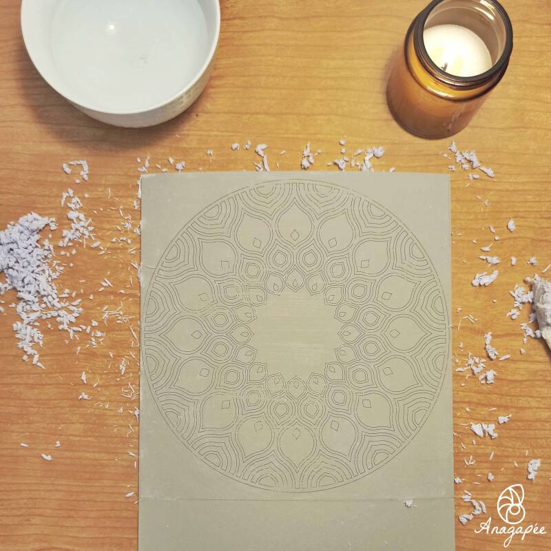 Une bougie pour la décoration, un petit bol contenant de l'eau, une plaque de Linoleum sur laquelle on voit le report d'un motif type mandala, et tout autour, des bouloches de papier, résidus de l'impression laser du motif une fois collé sur le lino et re