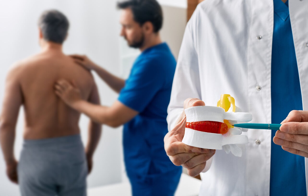 Das Bild zeigt einen Arzt, der ein Wirbelsäulenmodell hält, um Bandscheibenvorfälle zu erklären, während er einen Patienten mit Rückenschmerzen untersucht.