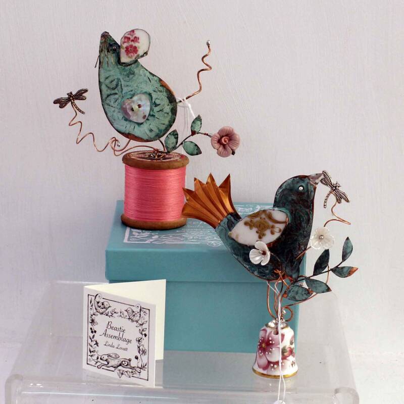 Two mini sculptures by artist Linda Lovatt - one featuring a verdigis copper mouse sitting on an old wood cotton reel with a tiny metal dragon fly and a flower on wire and the other featuring a copper bird sitting on top of a flowery thimble