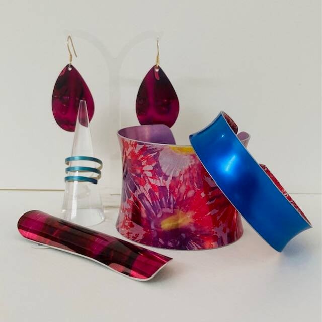 In shades of cerise. purple and blue, two wrist cuffs, a pair of tear shaped drop earrings, a spiral ring and a razor shell shape brooch all handmade in anodised alluminium by Miranda Peckitt of Very Colourful Jewellery