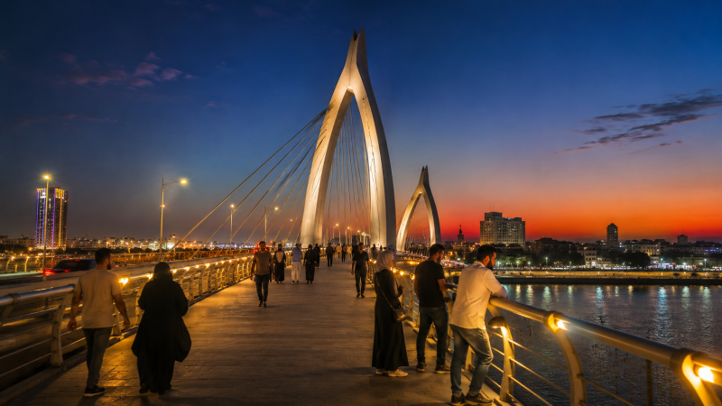 Wide evening view of the Dalal Bridge illuminated against a colorful sunset sky, with people walking, relaxing, and enjoying the atmosphere. The river reflects the city lights, creating a vibrant and peaceful scene that captures modern life and the beauty