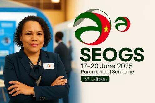 Zakelijke vrouw met een vriendelijke glimlach in een donkerblauw pak staat met gekruiste armen voor het SEOGS 2025-logo en eventinformatie. Op de achtergrond is een beursvloer te zien. De tekst vermeldt: “SEOGS, 17-20 June 2025, Paramaribo | Suriname, 5th