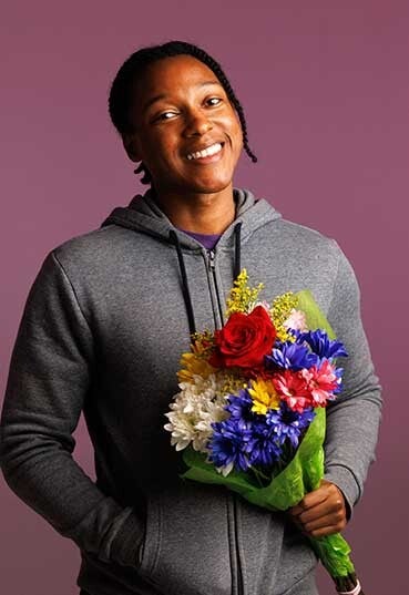 Purple background. Josh smiling wearing grey hoodie, holding mulicoloured flowers