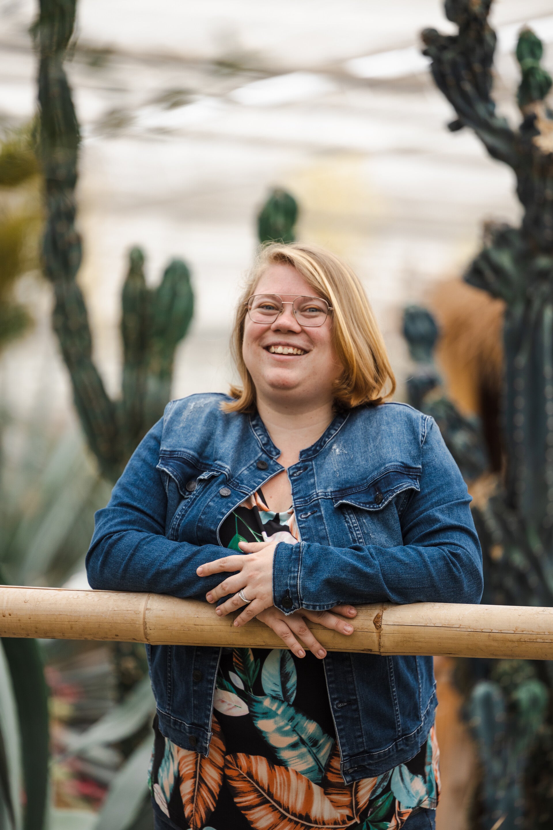 Een foto van Mara. Ze heeft blond haar en blauwe ogen en draagt een bril. Ze glimlacht breed. Op de achtergrond zijn cactussen te zien. Ze draagt een kleurrijk t-shirt en een spijkerjasje.