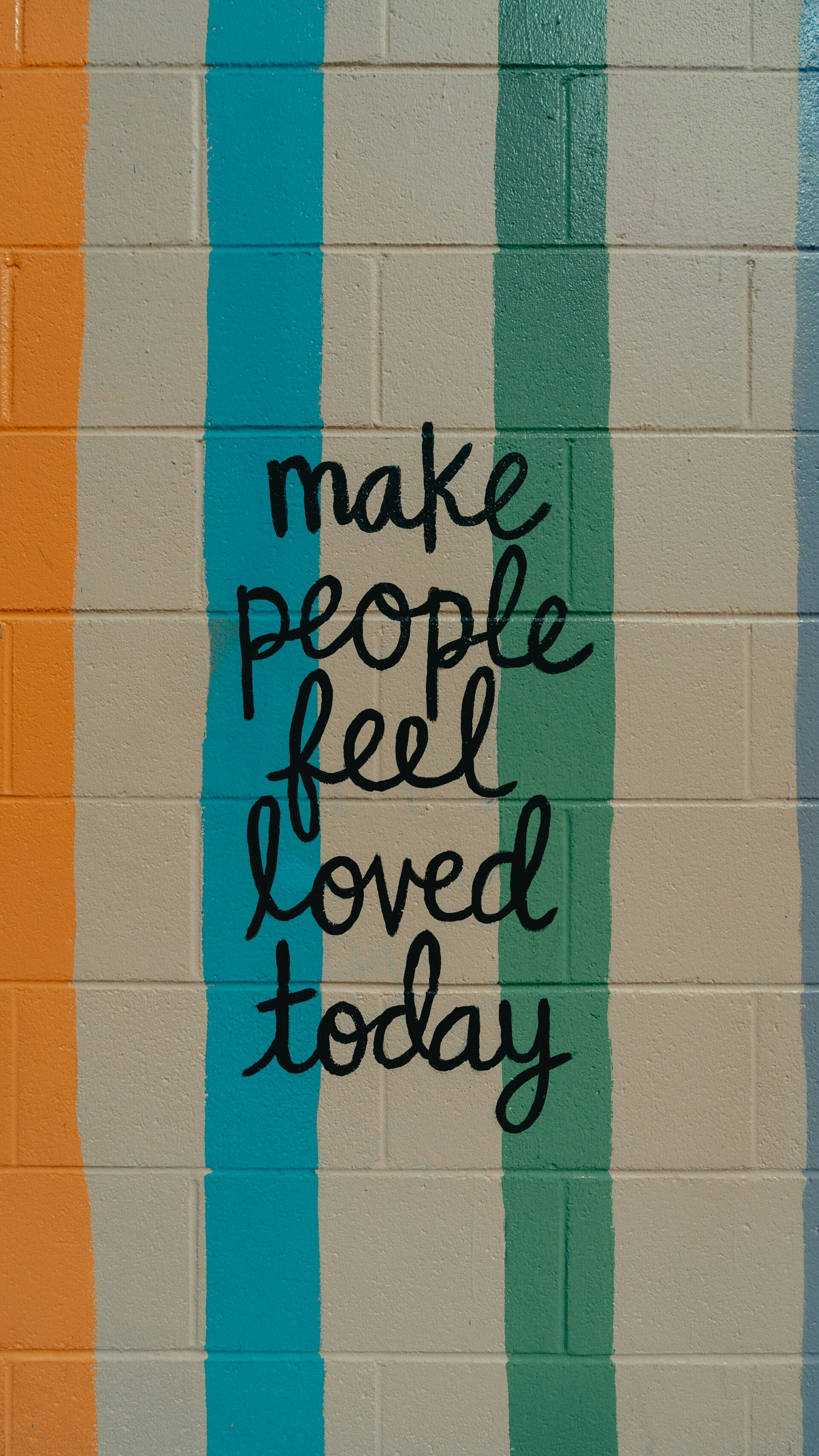 Een bakstenen muur met oranje, blauwe en groene strepen verf. In het midden staat geschreven: Make people feel loved today.