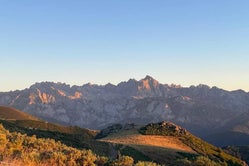 Paisaje natural de los Picos de Europa con laderas verdes, árboles y formaciones rocosas bajo un cielo azul despejado.