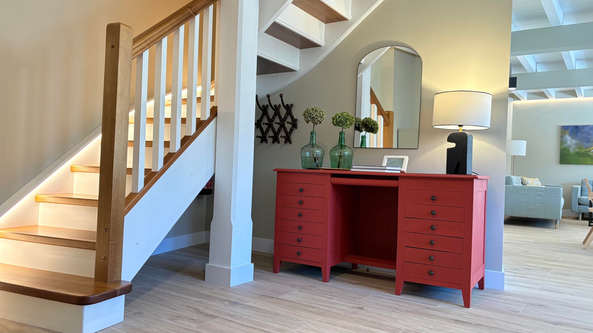 Interior de la casa rural Armaño 12 con escalera de madera iluminada, aparador rojo con lámpara y botellas decorativas, y un amplio salón comedor con mesa de madera, sillas trenzadas y sofá moderno.
