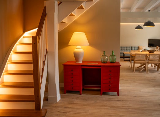 Interior de la casa rural Armaño 12 con escalera de madera iluminada, aparador rojo con lámpara y botellas decorativas, y un amplio salón comedor con mesa de madera, sillas trenzadas y sofá moderno.