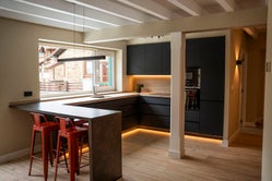 Cocina moderna en tonos oscuros con barra americana y taburetes rojos, iluminada con luz ambiental y vistas al exterior en Armaño 12.