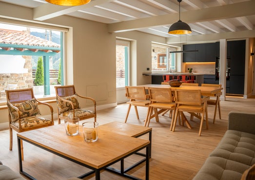 Amplio salón-comedor de la casa rural Armaño 12 con grandes ventanales, mesa de madera con sillas trenzadas, zona de estar con sillones y mesa de centro, y cocina abierta de diseño moderno al fondo.