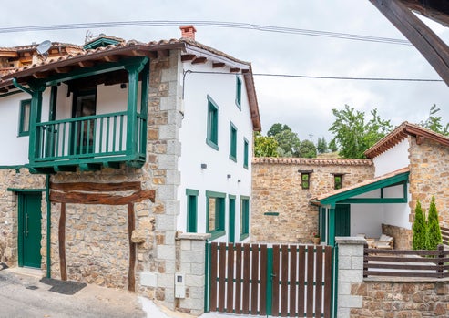 Fachada principal de la casa rural Armaño 12, con muros de piedra, balcones de madera verde y una entrada cercada, ubicada en un entorno rural de montaña en Liébana, Cantabria.