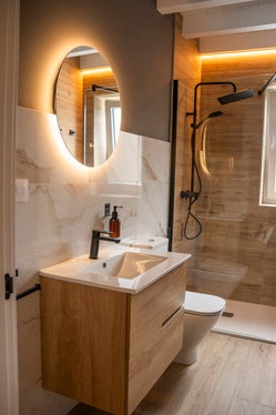 Baño moderno de Armaño 12 con ducha acristalada, mueble de madera, espejo redondo retroiluminado y grifería negra.