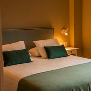 Habitación con cama doble, cabecero gris claro, cojines decorativos verdes y lámpara de lectura, en un ambiente relajante de la casa rural Armaño 12.
