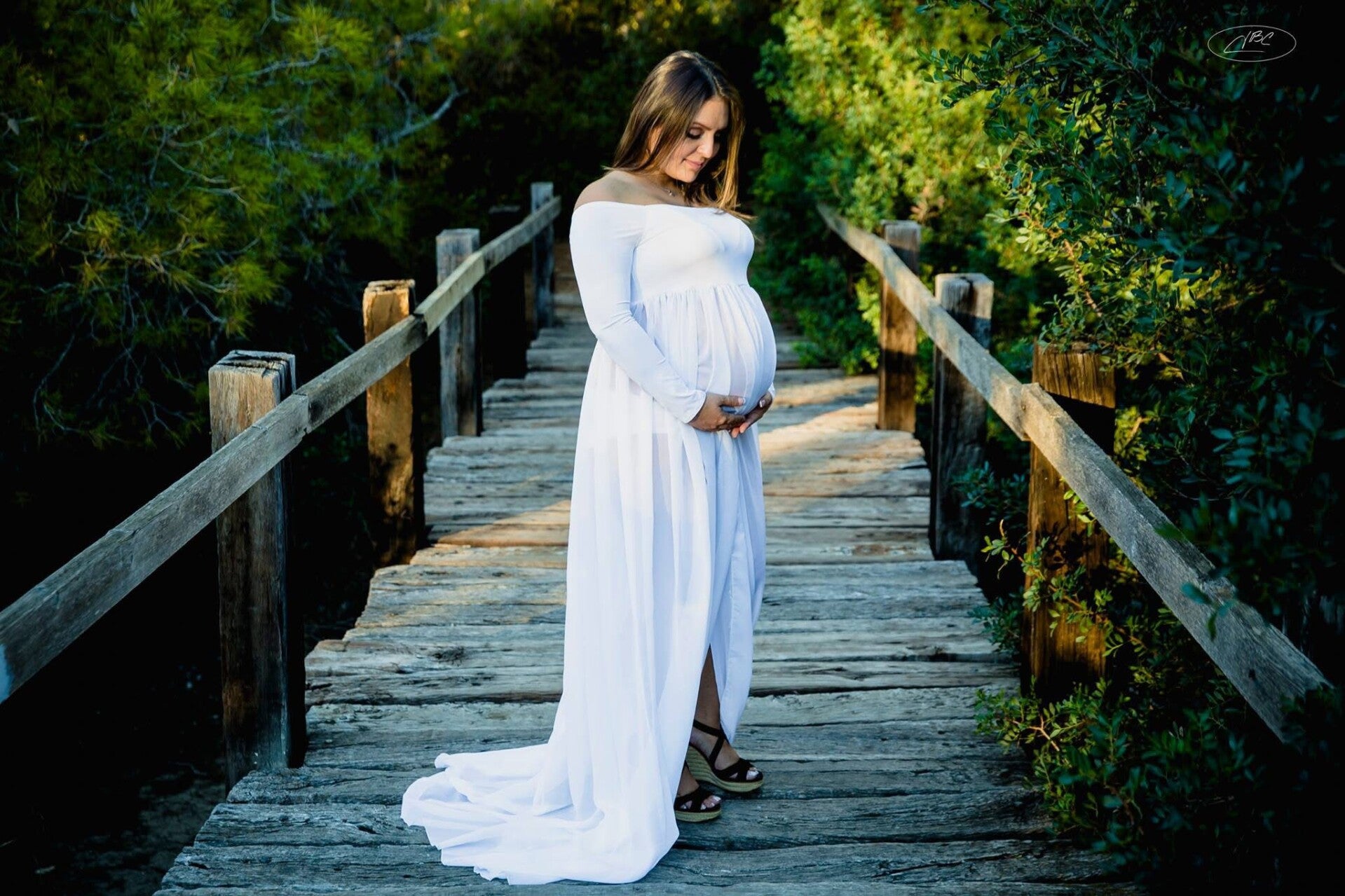 mujer embarazada caminando por un puente de madera en medio de un paisaje natural. Ella lleva un vestido blanco largo y fluido, que resalta su embarazo. Con una expresión suave, mira hacia su vientre mientras lo sostiene con ambas manos.