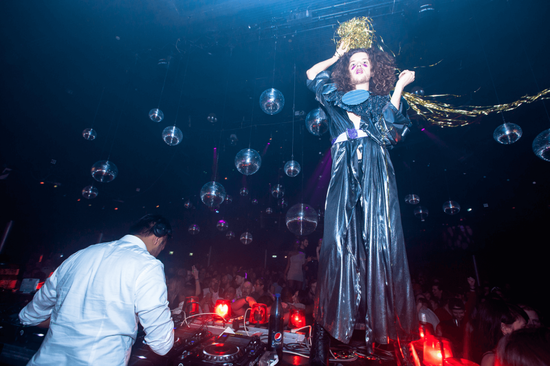 Man wearing silver dress, standing on a DJ Booth in a club