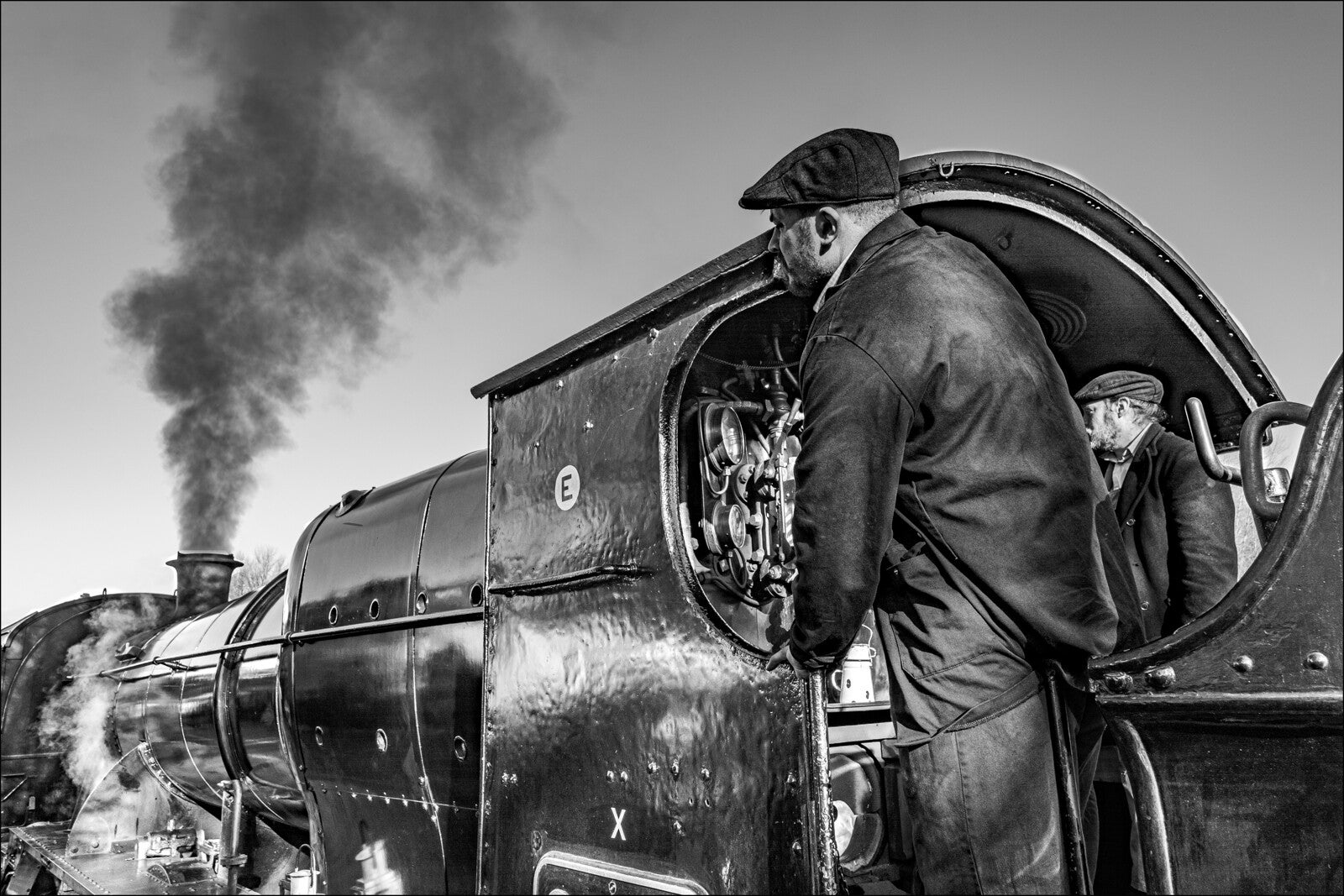Black and white photo of two crew members in caps inside the cab of a glossy steam locomotive. One man looks forward past the engine boiler, which releases a thick plume of dark smoke into the clear sky