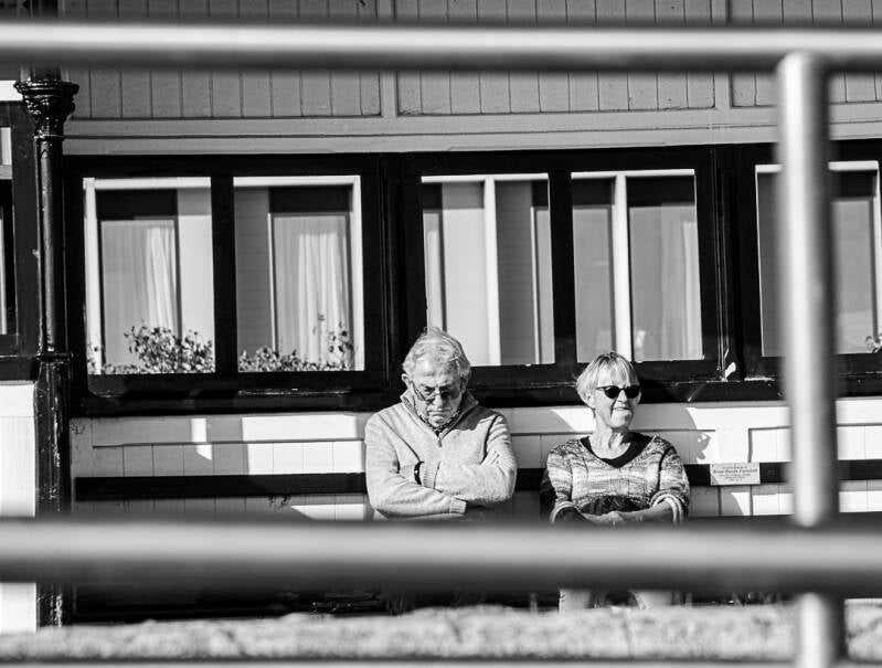 Candid black and white photograph of a couple sitting together by the seaside, framed by railings, with contrasting expressions and body language.