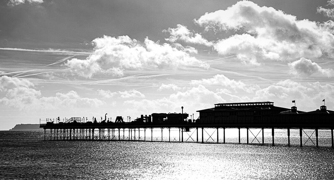 Black and white photograph of a seaside pier silhouetted against dramatic clouds, with sunlight reflecting across the water.