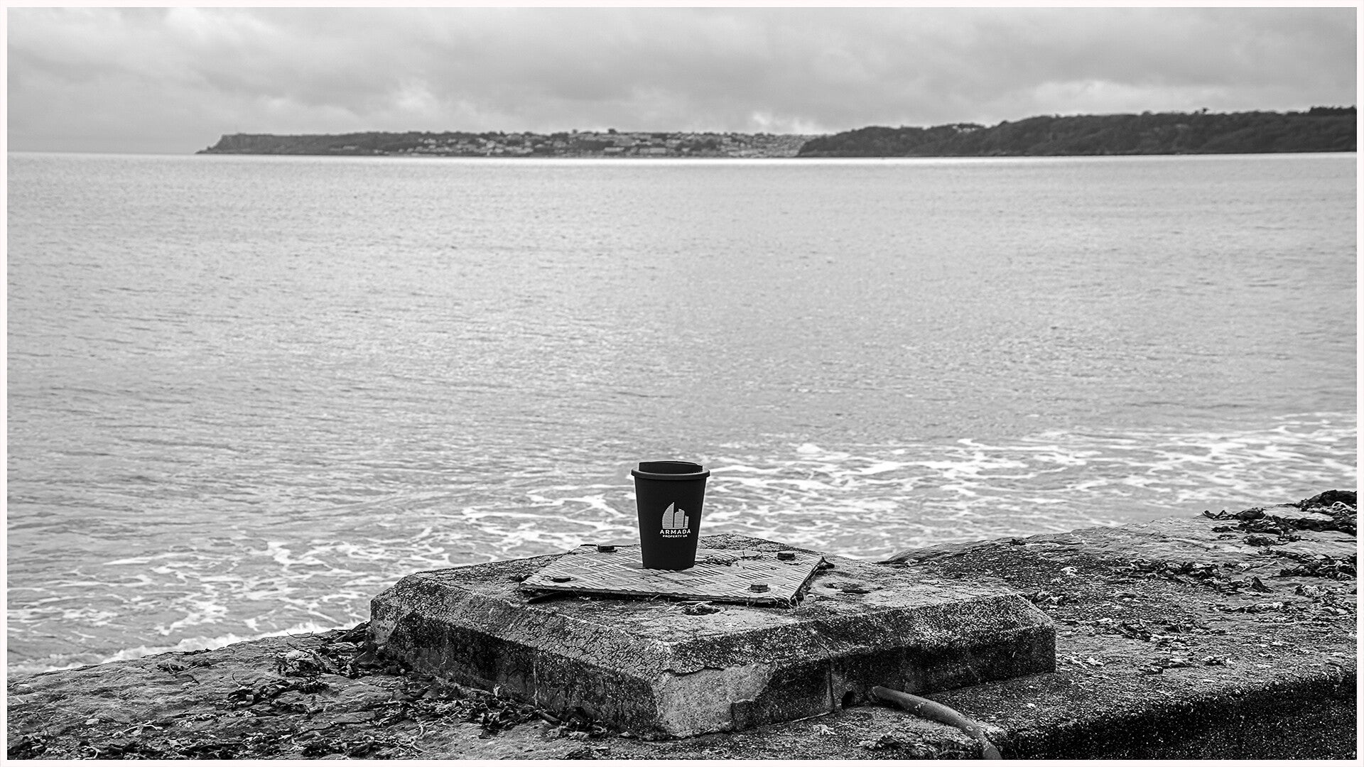 A solitary discarded takeaway coffee cup resting on a concrete platform overlooking the sea, with a distant coastline beneath a cloudy sky.