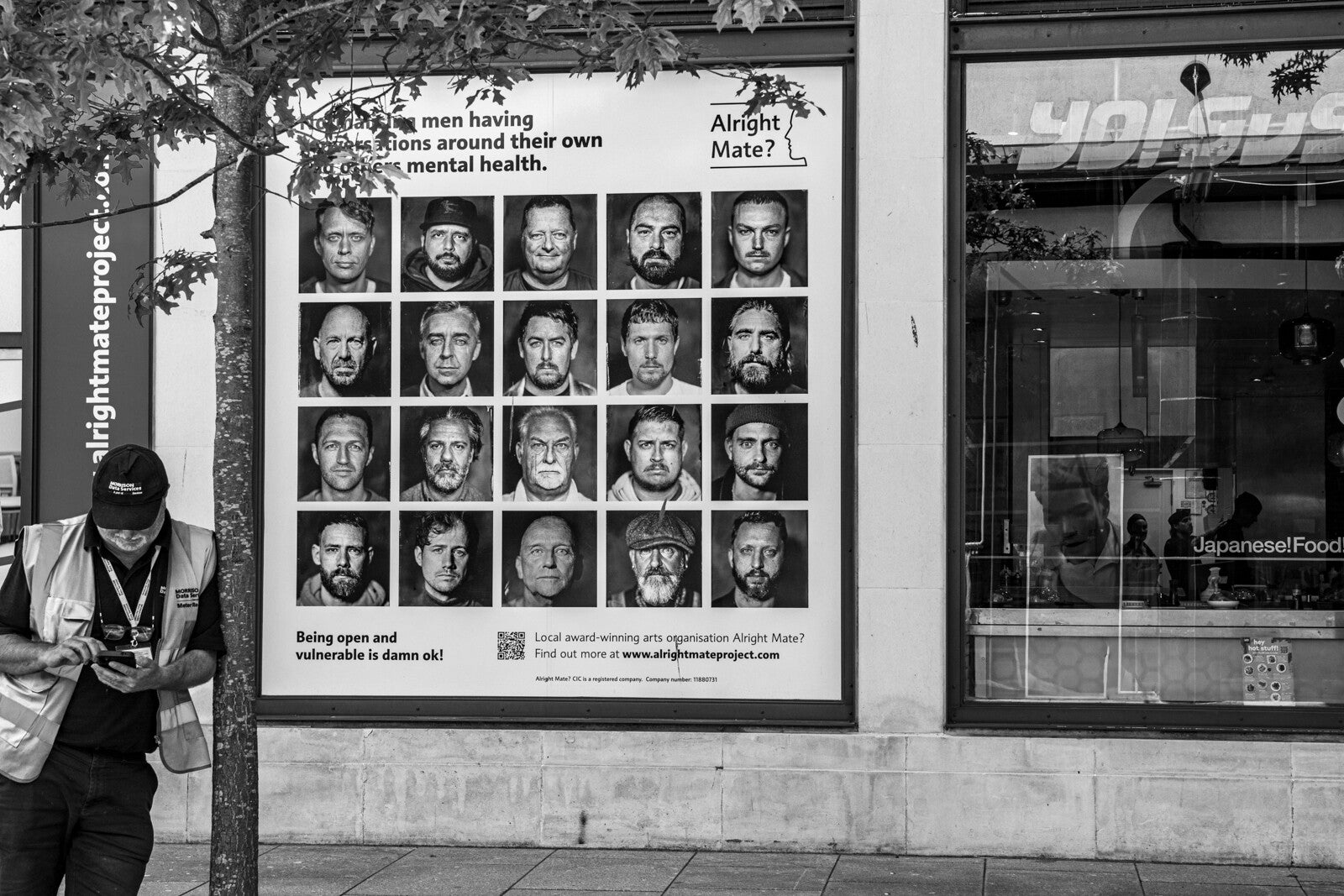 Black and white street photograph showing a man beside an Alright Mate mental health poster by Robbie Clymo