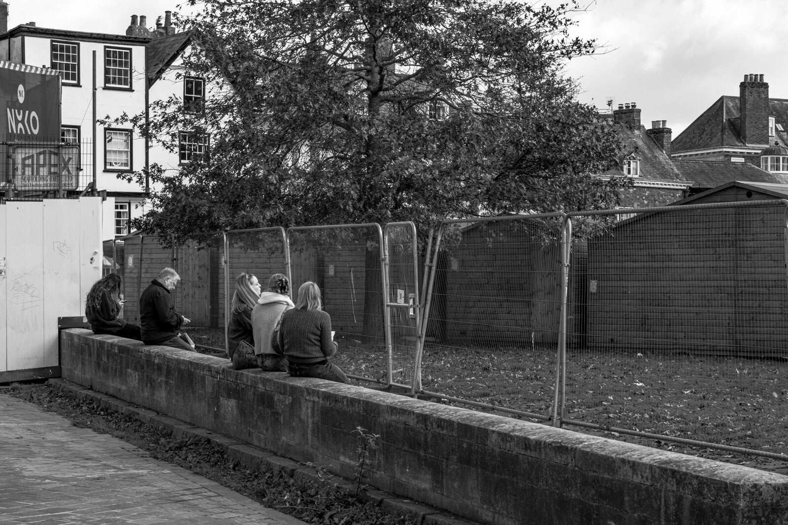 Black and white street photograph of people sitting beside construction fencing by Robbie Clymo