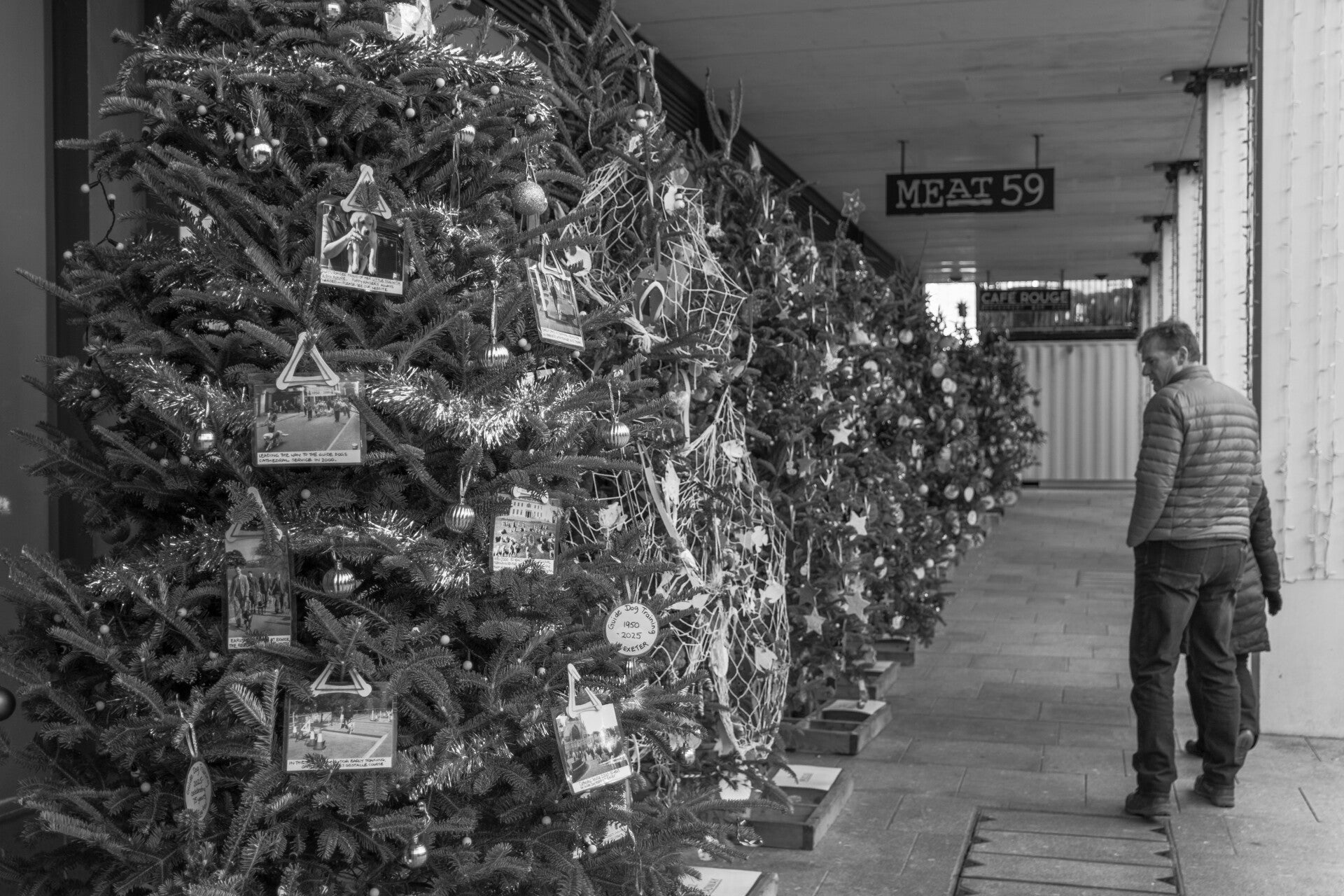 Black and white street photograph of a man walking past decorated Christmas trees by Robbie Clymo