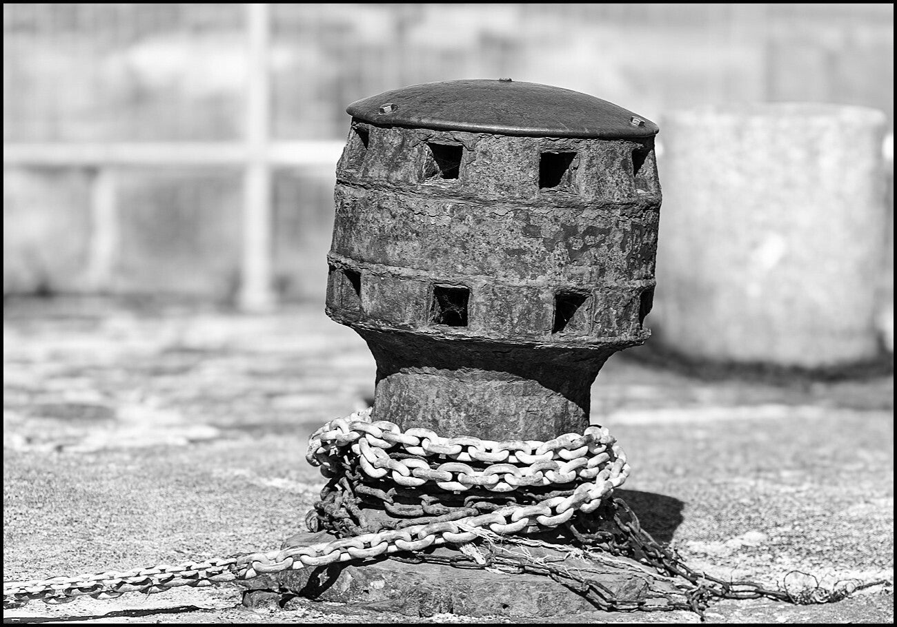 A black and white photograph focusing on a weathered, cast-iron harbor capstan or bollard. The cylindrical structure features a domed top and two tiers of square openings wrapped around its upper section. It has a heavily textured, rusted, and peeling.