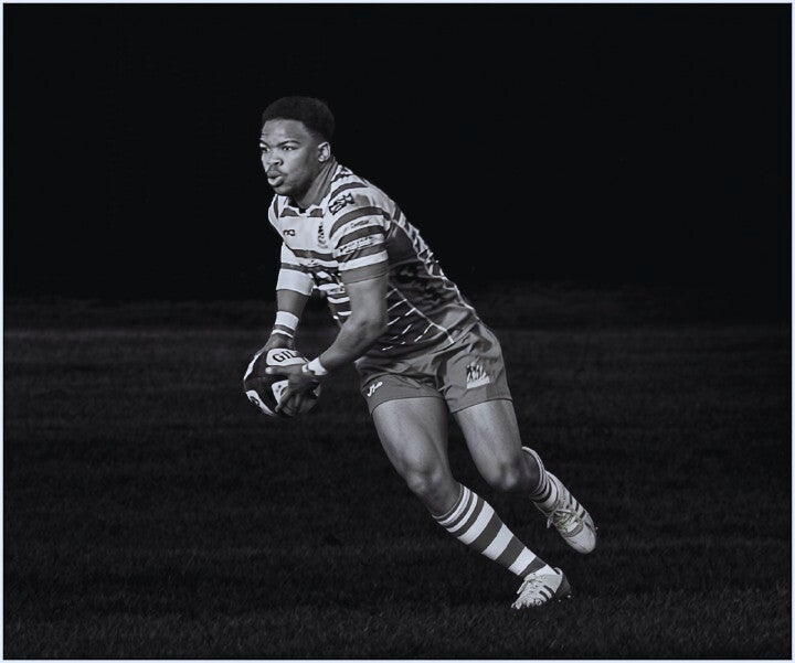 Black and white action photograph of a rugby player carrying the ball during a match.