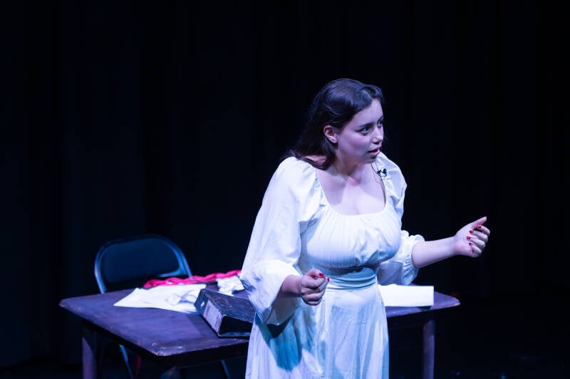 A woman in white standing in front of a messy desk. She is on stage.
