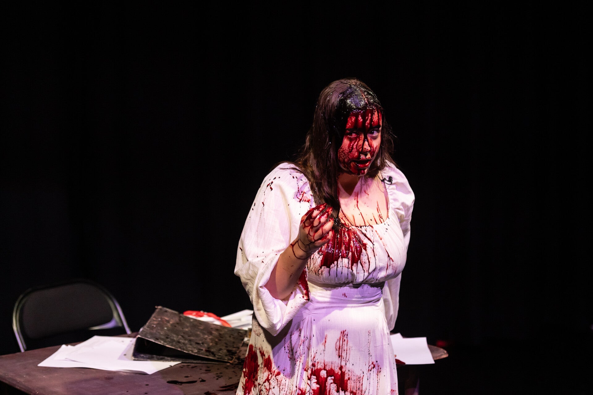 A woman in white standing in front of a messy desk. She is on stage.