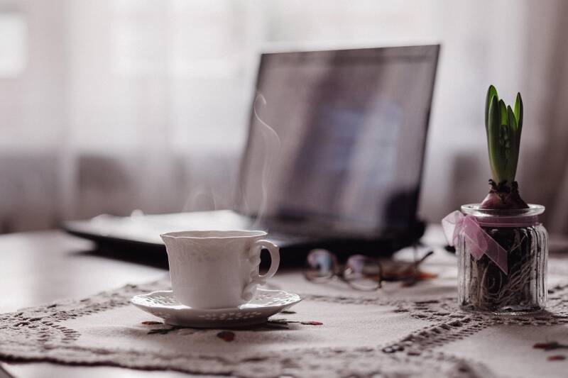 Dampfender Kaffeebecher auf Untertasse auf besticktem Tischtuch vor geöffnetem Laptop und blühender Pflanze in Glasgefäß, im Kontext mit Jenseitskontakte Jana Lorenz.