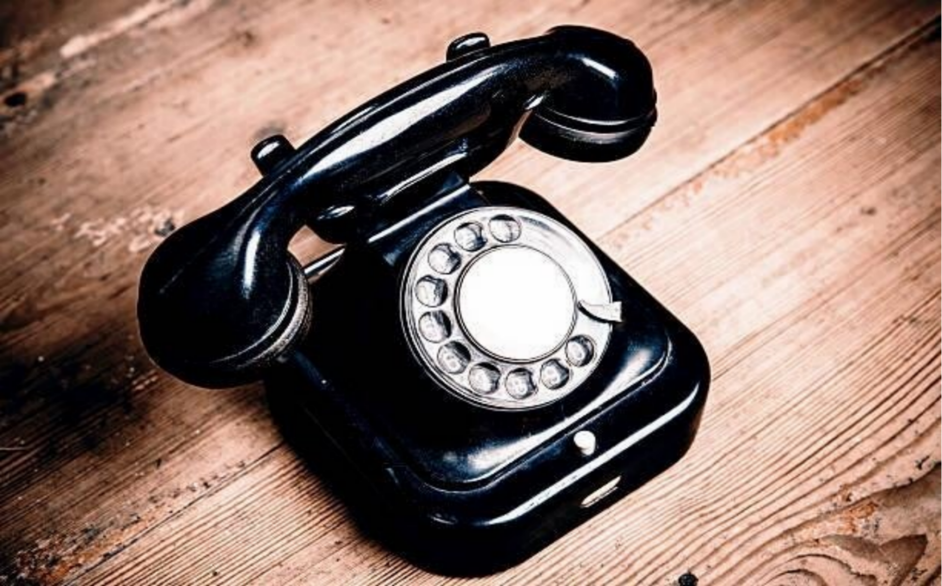 Ein klassisches, glänzend schwarzes Telefon mit Wählscheibe steht auf einem rustikalen Untergrund aus hellbraunen, strukturierten Holzdielen, im Kontext mit Jenseitskontakte Jana Lorenz.