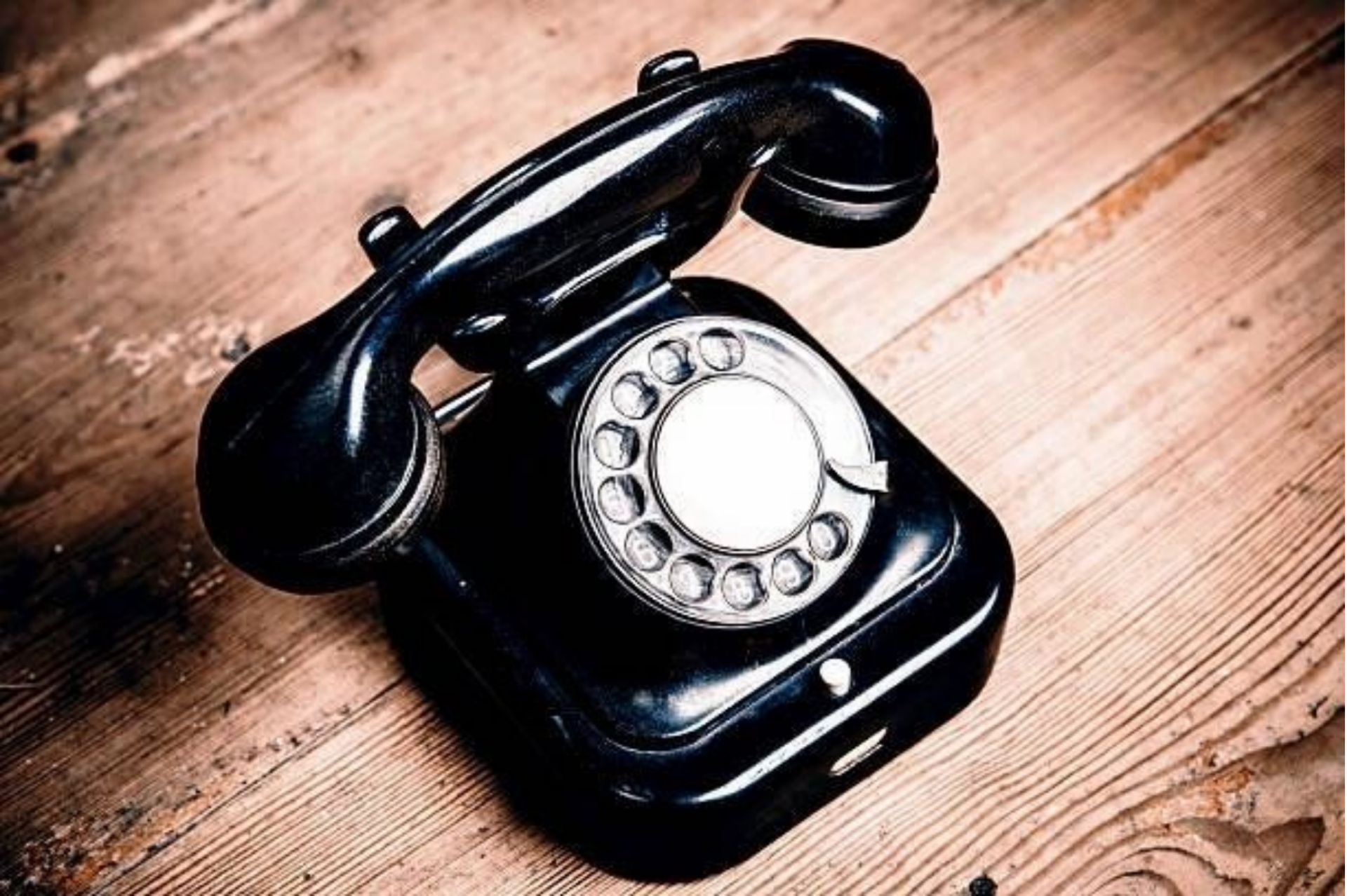 Ein klassisches, glänzend schwarzes Telefon mit Wählscheibe steht auf einem rustikalen Untergrund aus hellbraunen, strukturierten Holzdielen, im Kontext mit Jenseitskontakte Jana Lorenz.