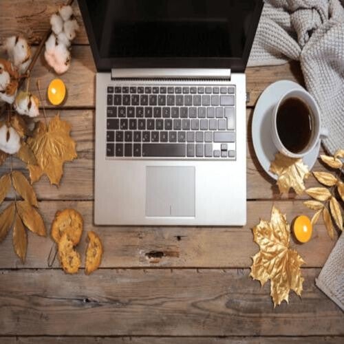 Ein Laptop auf einem Holztisch, umgeben von goldenen Herbstblättern, Baumwollzweigen, Keksen, einer Tasse Kaffee und einem grauen Strickpullover, im Kontext mit Jenseitskontakte Jana Lorenz.