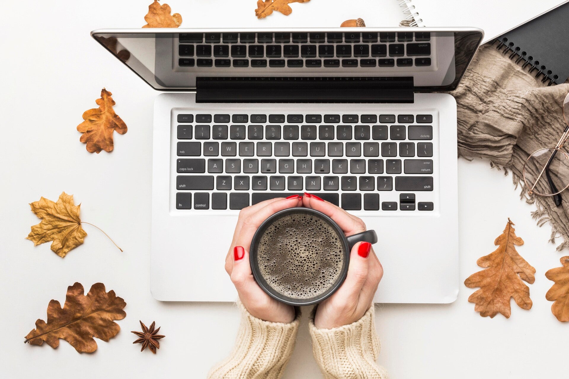 Hände mit rotem Nagellack halten eine Tasse Kaffee über einer geöffneten Laptop-Tastatur, umgeben von Herbstblättern und einem Notizbuch mit Stift, im Kontext mit Jenseitskontakte Jana Lorenz.