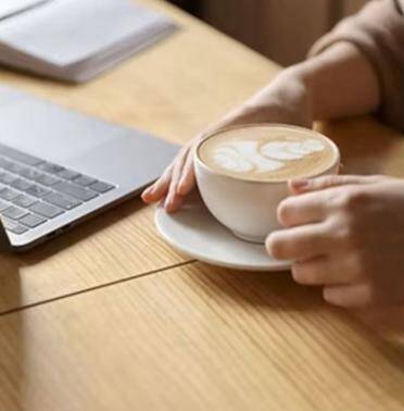 Hände halten eine Tasse Cappuccino mit Milchschaum, daneben ein aufgeklappter Laptop auf einem Holztisch, Kontext Kleine Gruppenlesung mit Medium Jana Lorenz