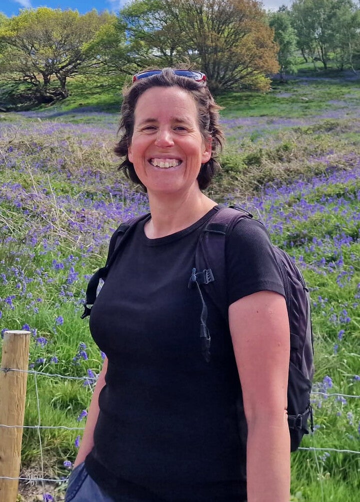 A medium close-up of Caroline giving her toothy smile in front of a field of bluebells, with her sunglasses on her head over medium length brown hair. She has a black backpack and t-shirt on. Photo by Alex Craig. 