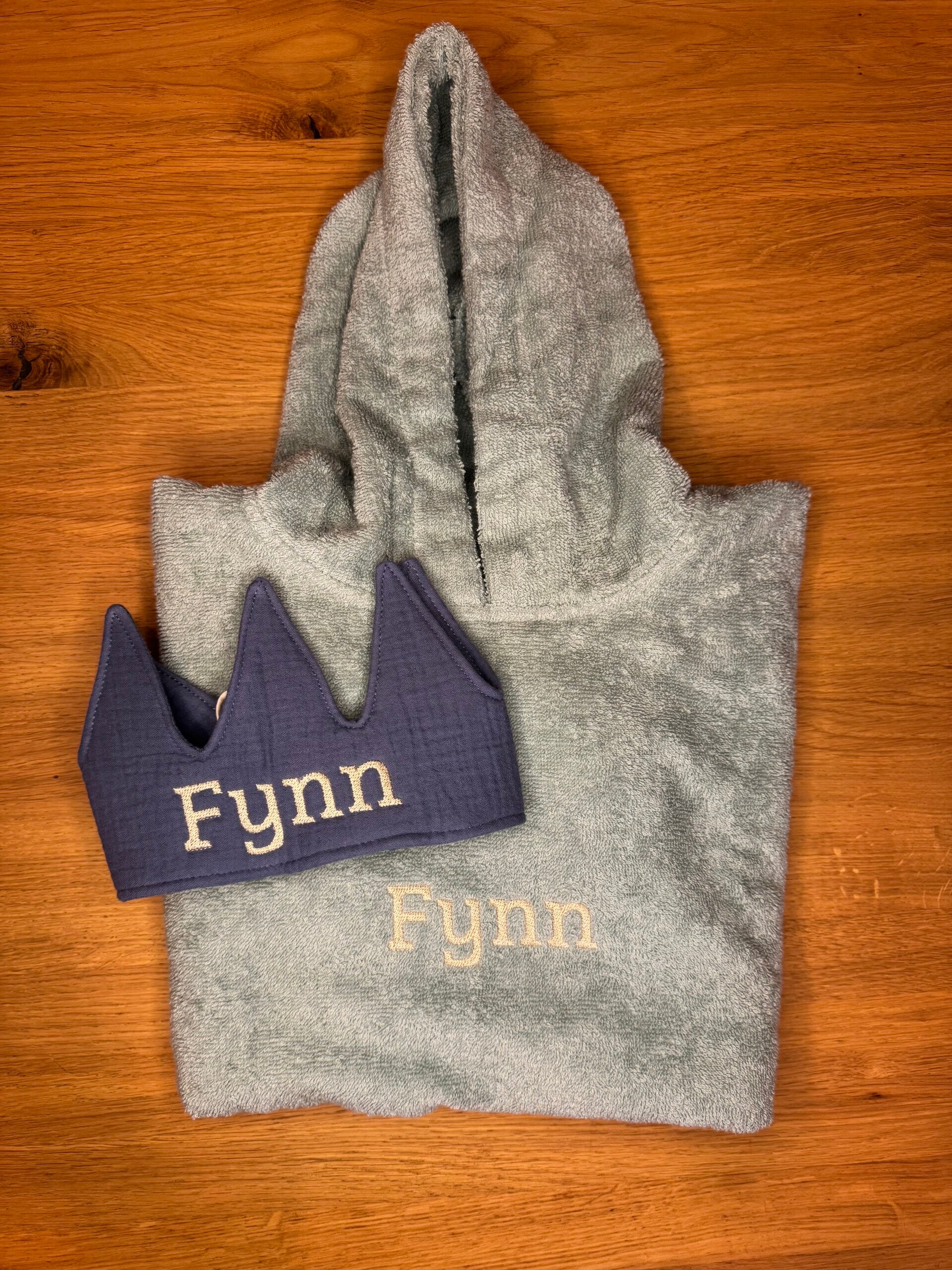 Een groene poncho met de naa Flynn met beige letters geborduurd en een jeans verjaardagskroontje met de naa Flynn in beige letters geborduurd