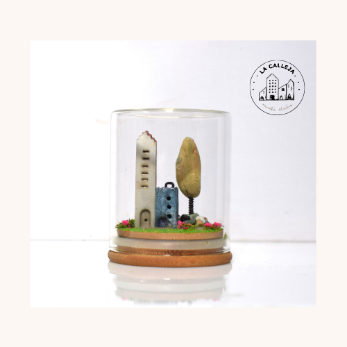 Escena encapsulada en cúpula de cristal con campanario blanco, casa azul y árbol en miniatura sobre base de madera.