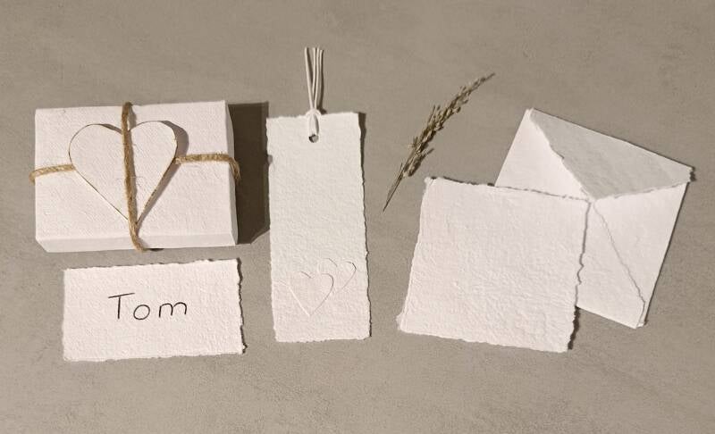 Weiße Schachtel aus handgeschöpten Papier mit Herz oben drauf gebunden mit Juteschnur weiße Namenskarte aus handgeschöpten Papier weißes Lesezeichen aus handgeschöpten Papier und Herz Applikation weißes Briefpapier aus handgeschöpten Papier 